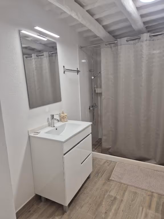 Casa de banho com lavatório branco, espelho, duche com cortina e tecto de vigas caiadas.