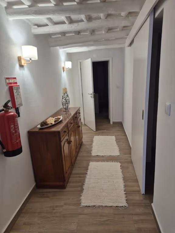 Corredor da casa com tecto de vigas caiadas de branco, uma cómoda antiga de madeira escura e dois apliques acesos.