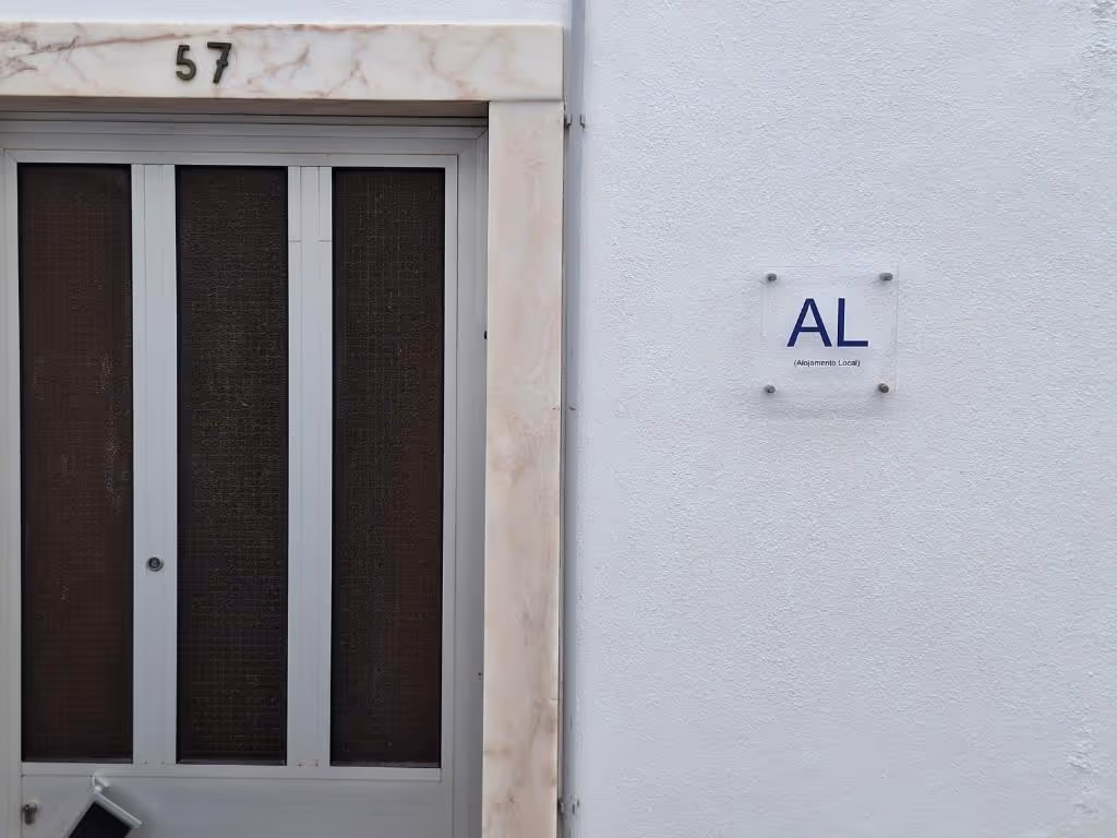 Porta de entrada da casa, com o número 57 em ferro sobre a padieira de mármore e a placa de Alojamento Local na parede caiada.