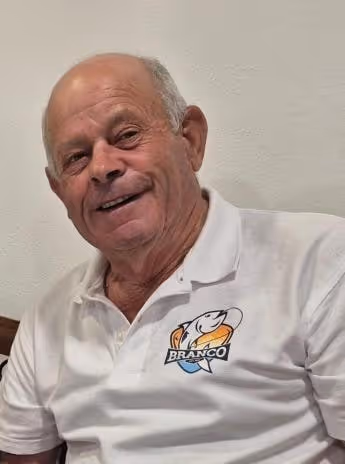 Joaquim Branco, sorridente, de camisa branca, sentado em casa.