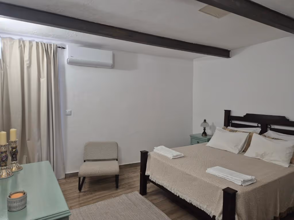 Quarto de casal com vigas escuras no tecto, ar condicionado, cama de madeira e cómoda verde-água.