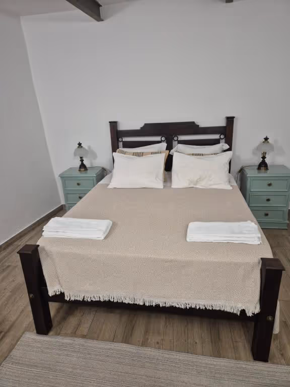 Quarto de casal com cama de madeira escura, duas mesas de cabeceira verde-água com candeeiros e toalhas dobradas sobre a colcha.