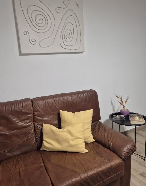 Sofá de pele castanha com uma almofada amarela e um quadro de traço na parede caiada.