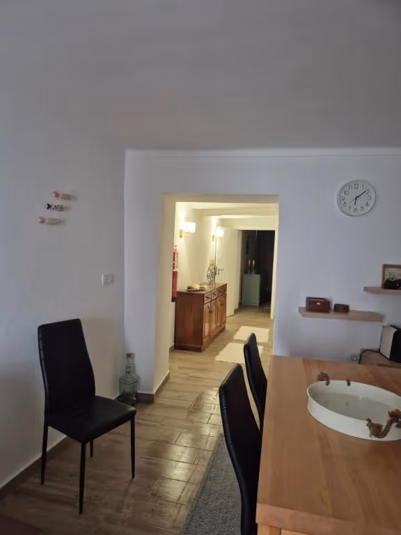 Sala de jantar com mesa de madeira, relógio de parede e três peixes de cerâmica pendurados, com o corredor ao fundo.
