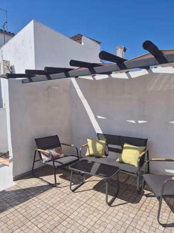 Terraço com pérgola de vigas escuras, cadeiras e mesa baixa, sobre chão de calçada, com céu limpo.